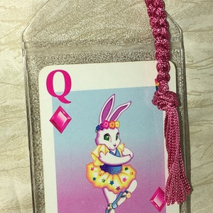 May include: A bookmark featuring a playing card design with a cartoon rabbit dressed as a ballerina. The card is the Queen of Diamonds, with a pink tassel and a clear plastic sleeve.