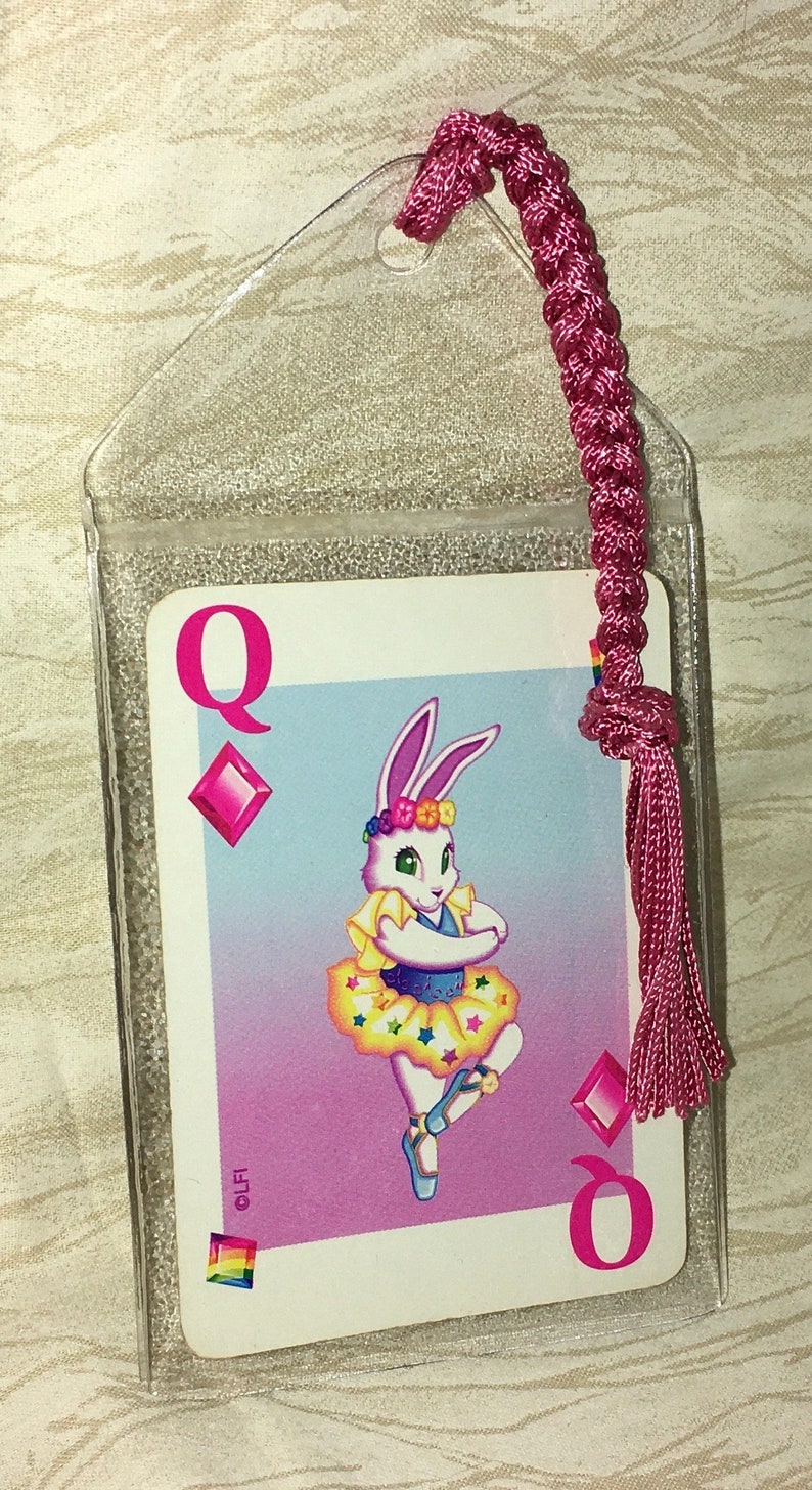 May include: A bookmark featuring a playing card design with a cartoon rabbit dressed as a ballerina. The card is the Queen of Diamonds, with a pink tassel and a clear plastic sleeve.