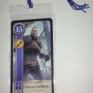 Peut inclure: Marque-page en plastique transparent avec une carte repr&eacute;sentant Geralt de Riv, personnage de la s&eacute;rie The Witcher. La carte a un design bleu et or avec le chiffre 15. Une corde tress&eacute;e bleue est attach&eacute;e en haut.
