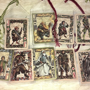 May include: Set of eight playing card bookmarks featuring characters from a fantasy series. Each card has a unique illustration and is encased in a clear plastic sleeve. The bookmarks are adorned with decorative cords in green and burgundy.
