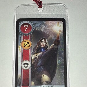 Peut inclure: Carte Yennefer de Vengerberg, repr&eacute;sentant une femme en cape sombre avec une main lumineuse. La carte a une bordure rouge et blanche avec des symboles et le chiffre 7. Un cordon bordeaux est attach&eacute; en haut.
