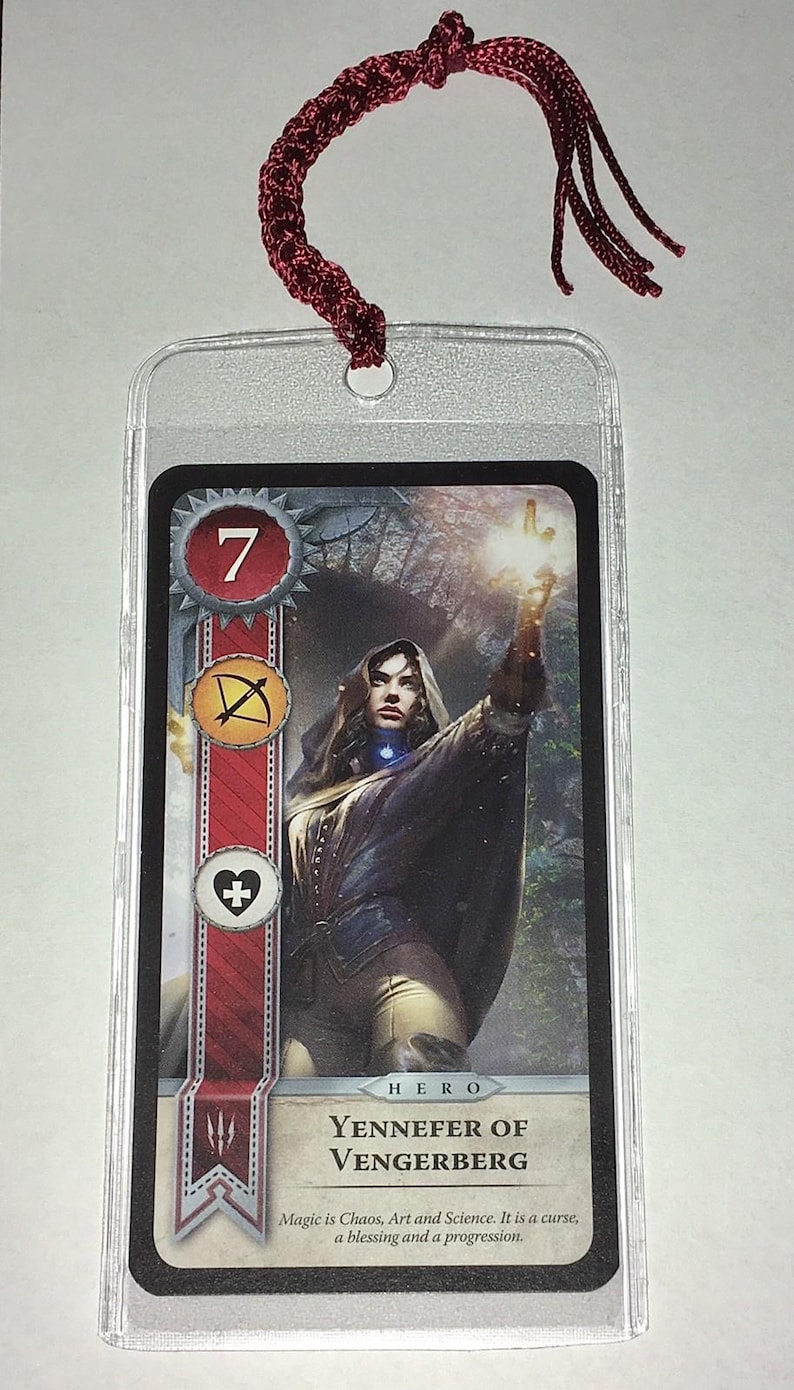 Peut inclure: Carte Yennefer de Vengerberg, repr&eacute;sentant une femme en cape sombre avec une main lumineuse. La carte a une bordure rouge et blanche avec des symboles et le chiffre 7. Un cordon bordeaux est attach&eacute; en haut.