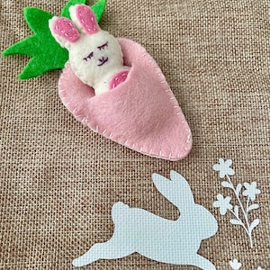 May include: A white felt bunny with pink ears is nestled inside a pink felt carrot. The carrot has a green felt leaf on top. A white felt bunny silhouette is hopping on a brown burlap background with white felt flowers and footprints.