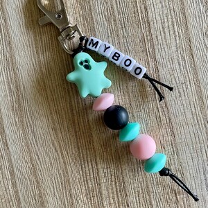 Op de afbeelding: Een sleutelhanger met een zilveren sluiting, een lichtblauwe spookbedel en de tekst "MY BOO" gespeld met witte letterkralen. De sleutelhanger heeft ook een reeks kleurrijke kralen in roze, zwart en teal.