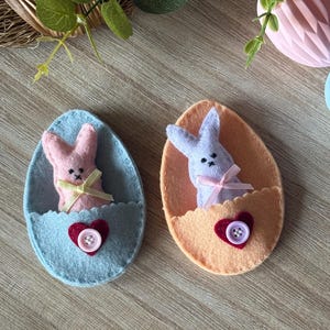 May include: Two felt Easter egg decorations. One is light blue with a pink bunny and a red heart with a button. The other is peach with a lavender bunny and a red heart with a button. Both have ribbon bows.