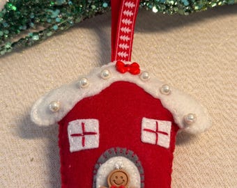 May include: A red felt Christmas ornament shaped like a house, with white accents and faux pearl embellishments. The house features white cross-shaped windows and a gingerbread man in the doorway. A red ribbon with a white pattern is attached for hanging.