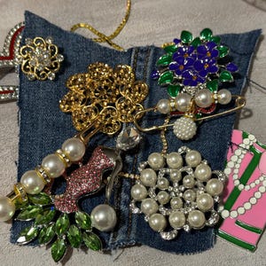 May include: A collection of assorted decorative pins and brooches featuring various designs including a gold-tone flower, a purple and green floral pin, a pearl-accented pin, a pink rose emblem, and a heart-shaped pin with red and white enamel.