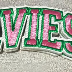 May include: A fabric patch featuring the word IVIES embroidered in pink thread with a bright green border and a white base layer. The letters are block-style and have a raised, textured appearance with a distinct stitched edge finish.