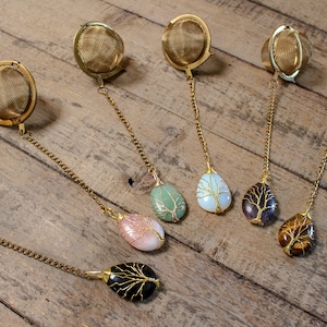 May include: Set of six gold tea infusers with different colored gemstone pendants. Each pendant features a wire tree design. The gemstones are green, white, purple, brown, pink, and black.