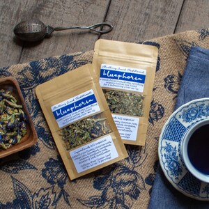 May include: An overhead shot features two pouches of "Bluephoria" herbal tea, a cup of blue tea, and a tea infuser. The pouches have labels with text. Dried blue flowers are in a wooden bowl. The tea is in a blue and white patterned teacup.