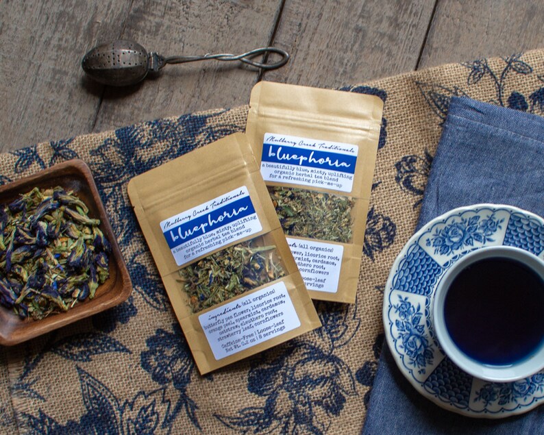 May include: An overhead shot features two pouches of "Bluephoria" herbal tea, a cup of blue tea, and a tea infuser. The pouches have labels with text. Dried blue flowers are in a wooden bowl. The tea is in a blue and white patterned teacup.