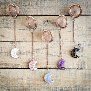 May include: Six copper tea infusers with different crystal moon charms. The infusers are hanging from a wooden background.