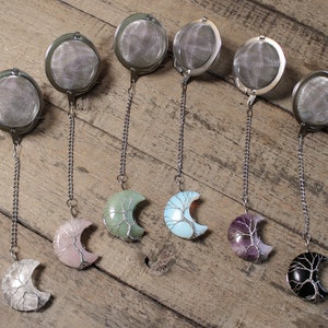May include: Seven silver tea infusers with chains and different colored crescent moon-shaped crystal charms. The crystals are green, blue, pink, purple, and black.