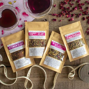 May include: Four small brown paper bags with clear windows showing different types of loose leaf tea blends. The bags are labelled with the names "Afternoon Delight", "Happy Heart", "Flower Garden Scents", and "Love Potion".