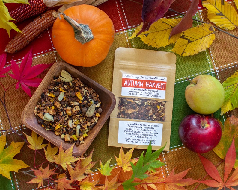 May include: An overhead shot of a fall-themed arrangement. A small orange pumpkin, a wooden bowl of tea, and a package of "Autumn Harvest" tea are surrounded by colorful leaves, apples, and corn. The tea package lists ingredients.