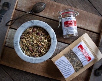 Può includere: Un vassoio di legno con una ciotola di marmo bianco piena di foglie di tè sfuse, un barattolo di vetro di Vital Tea Adaptogenic Tonic e un sacchetto di carta marrone con Vital Tea Adaptogenic Tonic scritto su di esso.