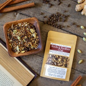 May include: A wooden bowl filled with chai tea blend, next to a package of Roots & Rooibos Chai. Cinnamon sticks, cardamom pods, and peppercorns are scattered on a wooden surface.