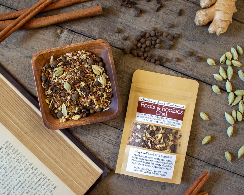 May include: A wooden bowl filled with chai tea blend, next to a package of Roots & Rooibos Chai. Cinnamon sticks, cardamom pods, and peppercorns are scattered on a wooden surface.