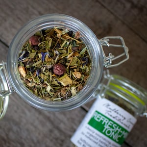 May include: Overhead shot of a clear glass jar filled with dried herbs, berries, and flowers. The jar has a metal clasp and a bright green rubber seal. A smaller jar with a label that reads "Refresh-Mint Tonic" is also visible.