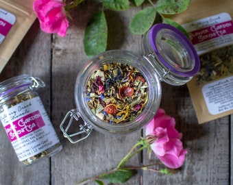 May include: A glass jar filled with a colorful loose-leaf herbal tea blend, with a purple-rimmed lid. The jar is open, revealing dried flowers and herbs. Pink rose blooms and green leaves surround the jar, with small tea packets visible.