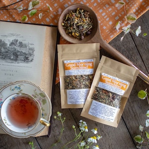 May include: An open book with a black and white illustration, a teacup with tea, and two small pouches of herbal tea blend. The pouches are labeled "Good Morning Sunshine!" and list ingredients. A wooden spoon holds loose tea.