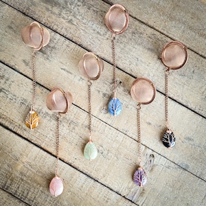 May include: Copper-colored tea infusers with chain and gemstone pendants. The infusers have a mesh basket design. Gemstone pendants include yellow, pink, green, blue, purple, and black, each with a tree of life design.