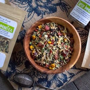 May include: A wooden bowl filled with a blend of dried herbs and flowers for Super Immunitea, a powerful immune-boosting organic herbal tea blend. The tea is caffeine-free and comes in a 0.7 oz bag, making 112 servings. The ingredients include echinacea, turmeric root, elderberries, rosehips, astragalus root, ginger root, nettle, cinnamon, licorice root, sage, lemon thyme, and calendula flowers.