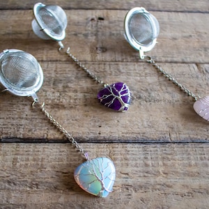 May include: Three silver tea infusers with heart-shaped crystal charms. The charms are amethyst, rose quartz, and opal. Each charm has a wire tree design.