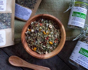 May include: A wooden bowl filled with a blend of dried herbs and spices, including turmeric, peppermint, and rose petals. Two glass jars labeled "AllerEase" with a description of the product are in the background. The jars are on a burlap surface.
