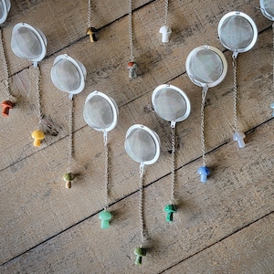 May include: A collection of 12 silver tea infusers with mushroom-shaped charms in various colors, hanging from a wooden surface.
