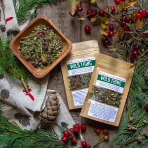 May include: An overhead shot features a wooden bowl filled with a herbal tea blend, alongside two pouches labeled "Wild Thing." The scene is decorated with pine branches, red berries, and a patterned cloth, creating a rustic, natural aesthetic.
