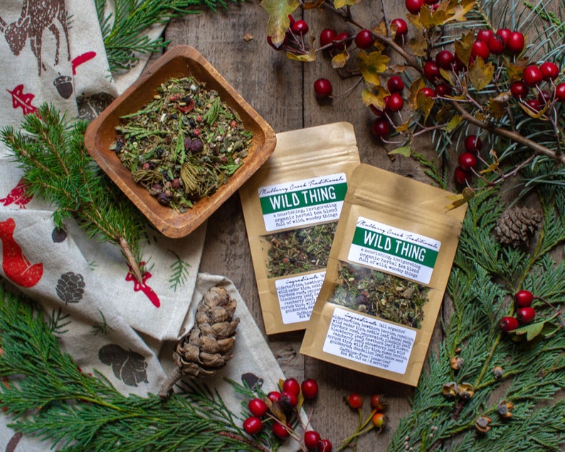 May include: An overhead shot features a wooden bowl filled with a herbal tea blend, alongside two pouches labeled "Wild Thing." The scene is decorated with pine branches, red berries, and a patterned cloth, creating a rustic, natural aesthetic.