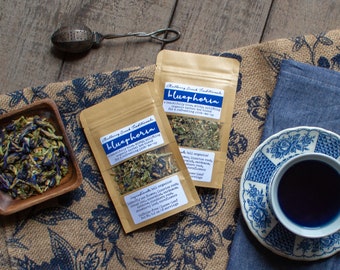 May include: Two brown paper bags of loose leaf tea labeled "Mallory Creek Traditional bluephoria" with a list of ingredients. The tea is a blend of organic herbs and flowers. A small bowl of blue and green tea leaves sits on a burlap table runner. A blue and white teacup with a dark blue tea sits on a matching saucer.