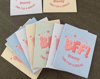 Op de afbeelding: Een stapel kleurrijke kaarten met de tekst "BFF! Stacey TIME FOR A BREAK" erop gedrukt. De kaarten zijn versierd met roze hartjes.