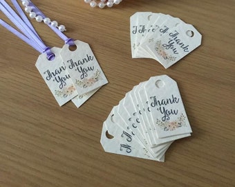Op de afbeelding: Een set van 12 witte cadeau-etiketten met een bloemenmotief en de tekst "Bedankt" in zwarte kalligrafie. De etiketten zijn vastgebonden met een lavendelkleurig lint en zijn in een waaier gerangschikt.