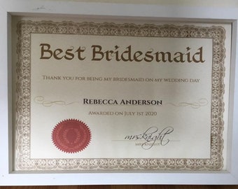 Op de afbeelding: Een ingelijst certificaat met een gouden rand en een rode zegel. De tekst luidt "Best Bridesmaid" en "Thank you for being my bridesmaid on my wedding day." Het certificaat wordt uitgereikt aan "Rebecca Anderson" op "1 juli 2020".