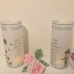 K&ouml;nnte beinhalten: Zwei wei&szlig;e, gefrostete Glaskerzenhalter mit gr&uuml;nen Kleeblatt-Designs und aufgedruckten Namen. Die Kerzenhalter sind mit gr&uuml;nen Kleebl&auml;ttern und dem Text "The Shamrock" und "Rechauns" verziert.
