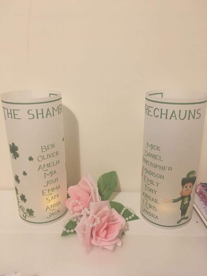 K&ouml;nnte beinhalten: Zwei wei&szlig;e, gefrostete Glaskerzenhalter mit gr&uuml;nen Kleeblatt-Designs und aufgedruckten Namen. Die Kerzenhalter sind mit gr&uuml;nen Kleebl&auml;ttern und dem Text "The Shamrock" und "Rechauns" verziert.