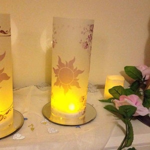 K&ouml;nnte beinhalten: Zwei gl&auml;serne Kerzenhalter mit floralem Design und einem Sonnenmotiv. Die Kerzen sind angez&uuml;ndet und verbreiten ein warmes Licht. Die Kerzenhalter stehen auf einer wei&szlig;en Oberfl&auml;che mit silbernen Glitzerherzen.