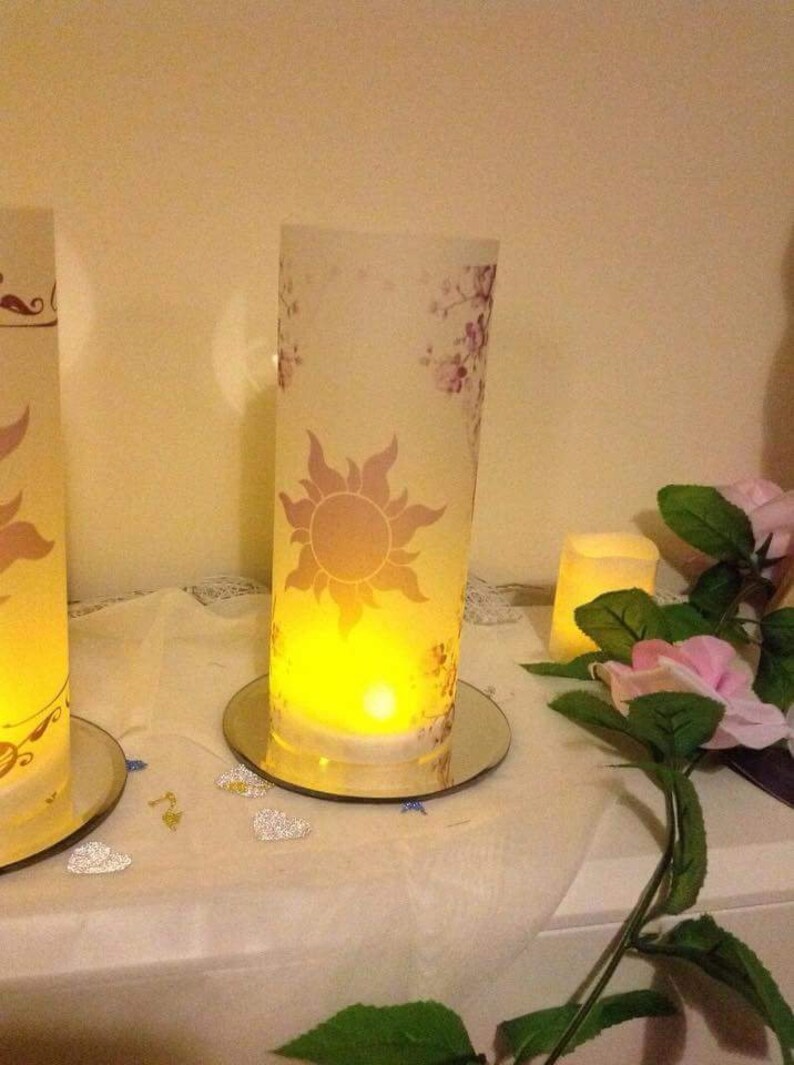 K&ouml;nnte beinhalten: Zwei gl&auml;serne Kerzenhalter mit floralem Design und einem Sonnenmotiv. Die Kerzen sind angez&uuml;ndet und verbreiten ein warmes Licht. Die Kerzenhalter stehen auf einer wei&szlig;en Oberfl&auml;che mit silbernen Glitzerherzen.