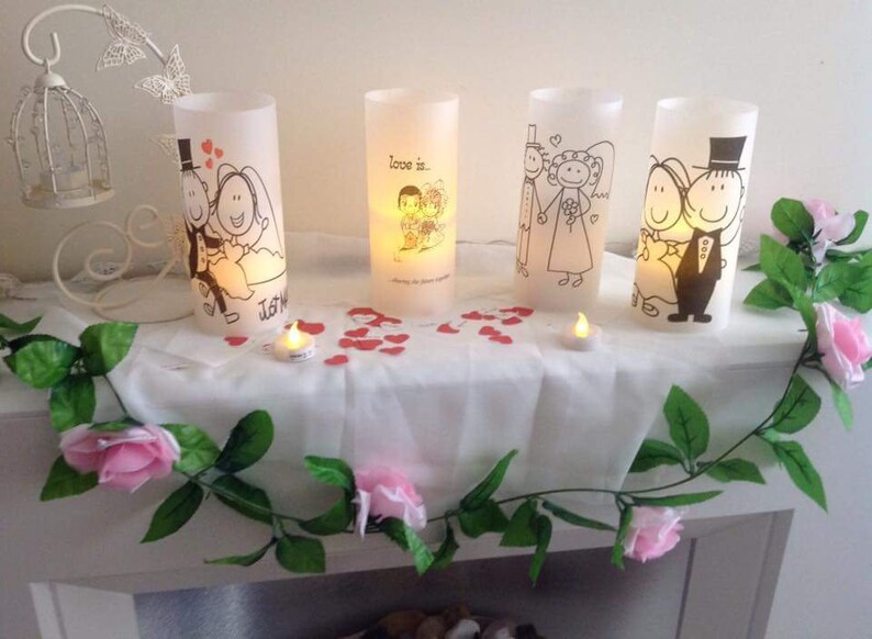K&ouml;nnte beinhalten: Vier wei&szlig;e zylindrische Kerzenhalter mit Illustrationen von Cartoon-Paaren. Die Kerzen sind angez&uuml;ndet und es sind rosa Rosen und rote Herzen um sie herum verstreut.