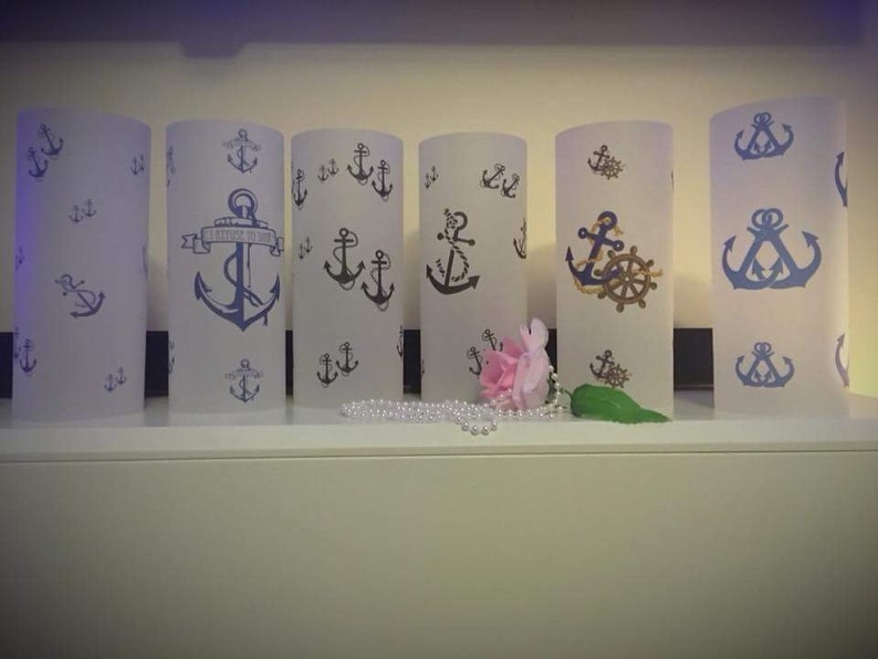 K&ouml;nnte beinhalten: Sechs wei&szlig;e, gefrostete Glaszylinder-Kerzenhalter mit blauen und goldenen nautischen Ankermotiven. Die Kerzenhalter sind in einer Reihe auf einem wei&szlig;en Regal angeordnet. Eine rosa Blume und eine Perlenkette befinden sich vor den Kerzenhaltern.