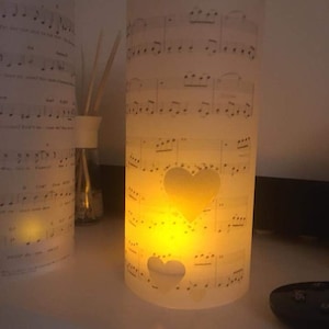 K&ouml;nnte beinhalten: Ein wei&szlig;er zylindrischer Kerzenhalter mit einem Design aus Noten und zwei Herz-Ausschnitten. Der Kerzenhalter ist angez&uuml;ndet und das Licht scheint durch die Ausschnitte und erzeugt ein warmes Leuchten. Der Text "Beauty and the Beast" ist oben auf dem Kerzenhalter sichtbar.