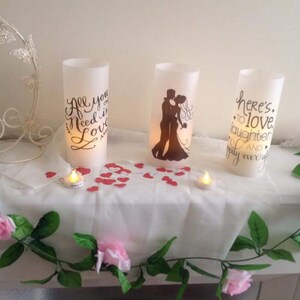 K&ouml;nnte beinhalten: Drei wei&szlig;e Kerzen in S&auml;ulenform mit unterschiedlichen Designs. Die Kerze links sagt "Alles was du brauchst ist Liebe". Die Kerze in der Mitte zeigt eine Silhouette eines Paares. Die Kerze rechts sagt "Hier ist auf Liebe, Lachen und ein gl&uuml;ckliches Leben". Die Kerzen stehen auf einem wei&szlig;en Tisch mit einer wei&szlig;en Tischdecke und rosa Blumen.