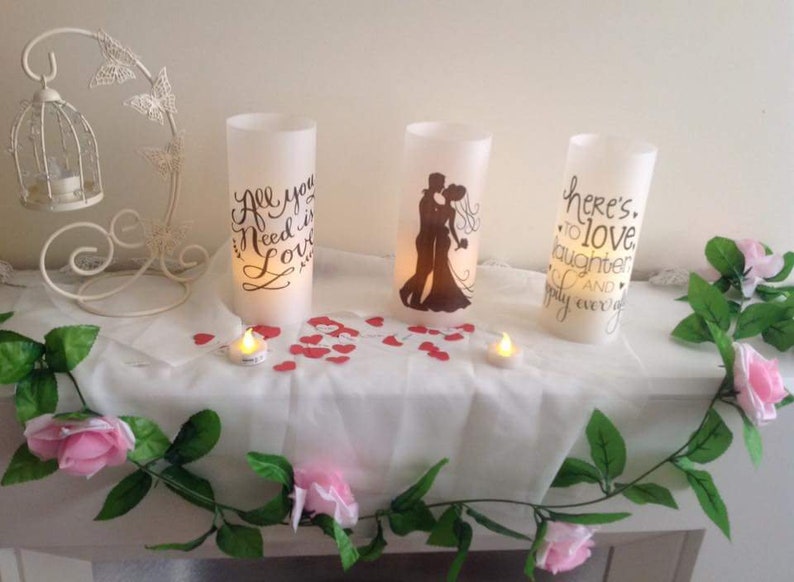 K&ouml;nnte beinhalten: Drei wei&szlig;e Kerzen in S&auml;ulenform mit unterschiedlichen Designs. Die Kerze links sagt "Alles was du brauchst ist Liebe". Die Kerze in der Mitte zeigt eine Silhouette eines Paares. Die Kerze rechts sagt "Hier ist auf Liebe, Lachen und ein gl&uuml;ckliches Leben". Die Kerzen stehen auf einem wei&szlig;en Tisch mit einer wei&szlig;en Tischdecke und rosa Blumen.