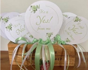 Op de afbeelding: Een set witte en groene ronde kaarten met de tekst "It's a Yes! from me" en "It's a No! from me" en "I said Yes! to the dress". De kaarten zijn gebonden met witte en groene linten en geplaatst in een houten doos.