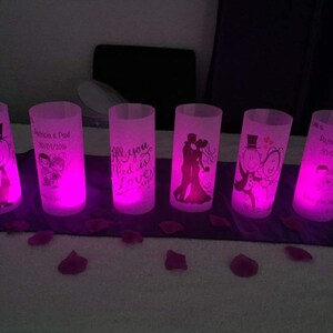 K&ouml;nnte beinhalten: Sechs klare Glaszylinder-Votivkerzenhalter mit rosa LED-Lichtern. Jeder Halter hat ein anderes Design, darunter ein Paar, eine Hochzeitstorte und den Text "Alles was du brauchst ist Liebe".