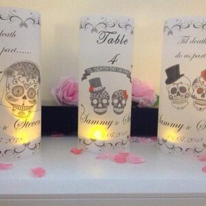 K&ouml;nnte beinhalten: Drei wei&szlig;e Papierlaternen mit einem schwarzen und wei&szlig;en Zucker Sch&auml;del Design. Die Laternen sind mit dem Text "Til death do us part..." und "Sammy & Steven" mit dem Datum "01.07.2018" verziert. Die mittlere Laterne ist eine Tischnummern-Laterne mit dem Text "Table 4" und "Til Death Do Us Part".