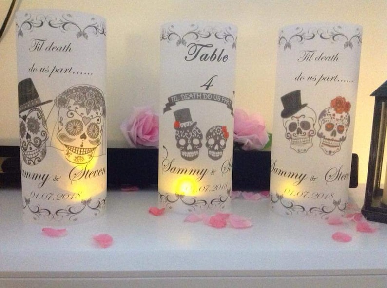 K&ouml;nnte beinhalten: Drei wei&szlig;e Papierlaternen mit einem schwarzen und wei&szlig;en Zucker Sch&auml;del Design. Die Laternen sind mit dem Text "Til death do us part..." und "Sammy & Steven" mit dem Datum "01.07.2018" verziert. Die mittlere Laterne ist eine Tischnummern-Laterne mit dem Text "Table 4" und "Til Death Do Us Part".