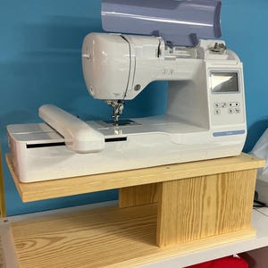 May include: A white sewing machine with a light blue cover sits on a light wood table. The machine has a digital display and various buttons. The table is supported by a lower shelf, also made of light wood.